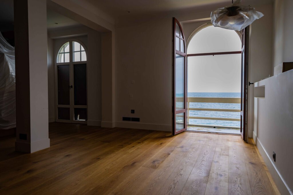 Pièce vide au parquet en bois avec porte voûtée ouvrant sur un balcon avec vue sur mer et rambarde de protection